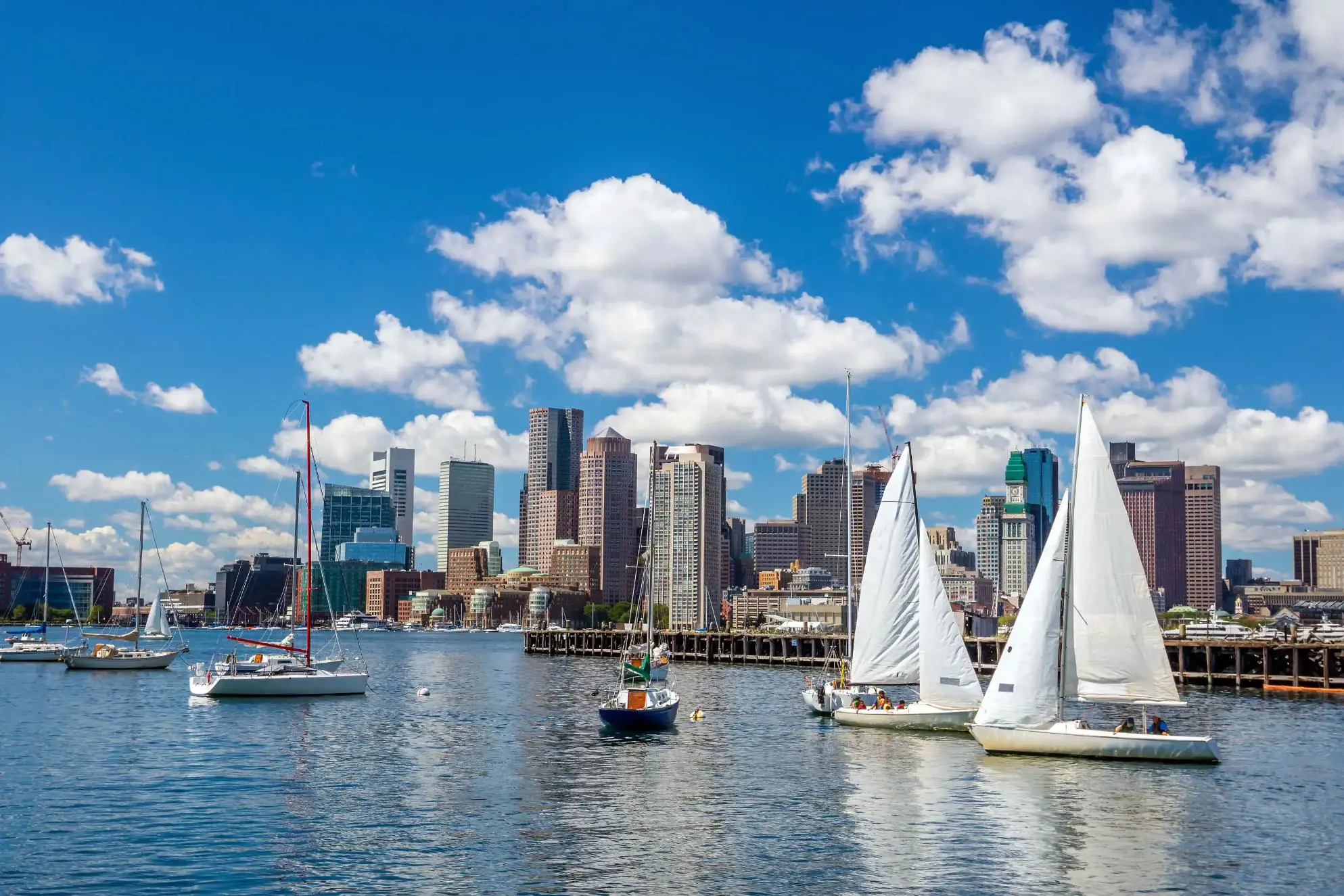 View of Boston, Massachusetts, seen across the water from Pier Park, with the city skyline rising above the harbour under a partly cloudy sky