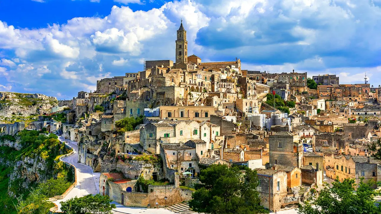 A panoramic view of the historic stone town of Matera, Italy, with ancient buildings stacked on a hillside under a partly cloudy blue sky