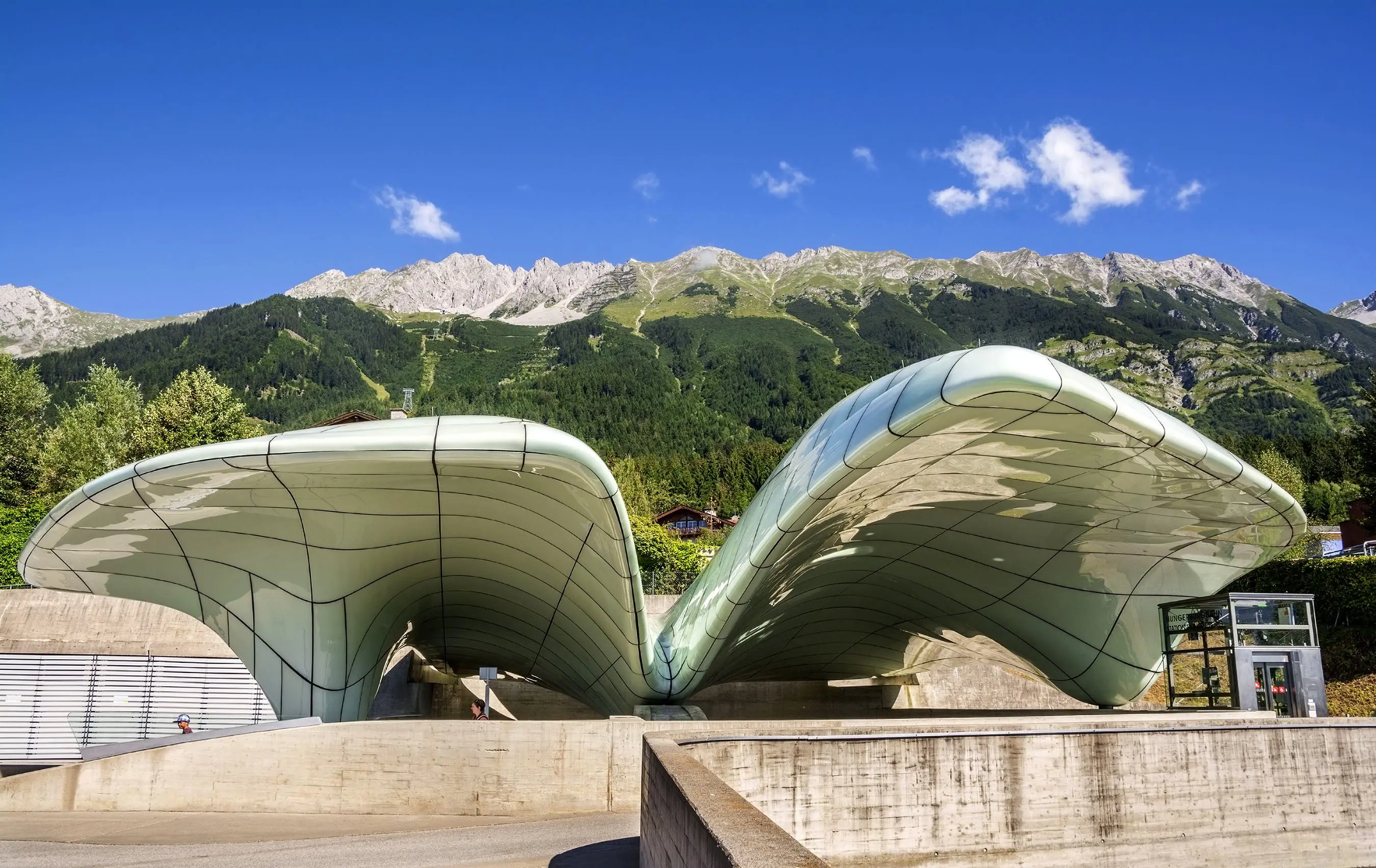 A white curved, horizontal, architectural structure with a thin, black cross pattern, in front of a grassy mountain with a clear blue sky behind it. Stone walls in the forefront 