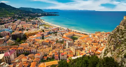 A panoramic view of Cefalù, Sicily, featuring its historic seaside town with terracotta rooftops, a sandy beach, and the rugged coastline beneath a lightly cloudy sky