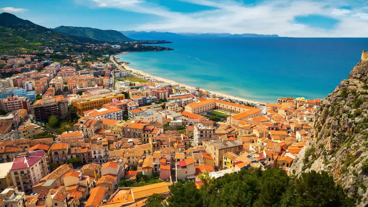 A panoramic view of Cefalù, Sicily, featuring its historic seaside town with terracotta rooftops, a sandy beach, and the rugged coastline beneath a lightly cloudy sky