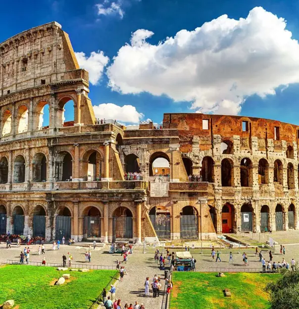The Colosseum in Rome, a vast ancient Roman amphitheatre with partially preserved stone arches and tiered seating, set against a blue sky with scattered clouds