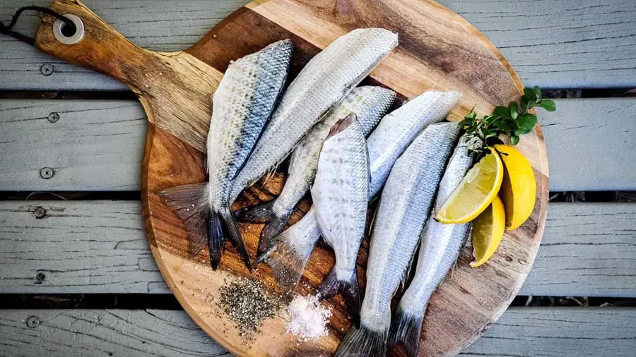 Whole silver fish on a wooden platter, with lemon slices, herbs and piles of salt & pepper next to them