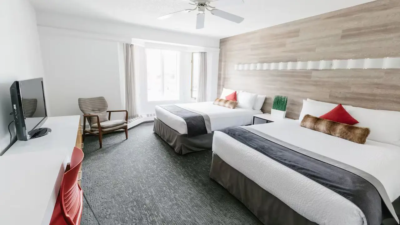 Spacious guestroom at Elk + Avenue hotel with two double beds, white bedding, grey bed skirts, a ceiling fan, a desk with a red chair, a flat-screen TV, and large windows allowing natural light to flood in