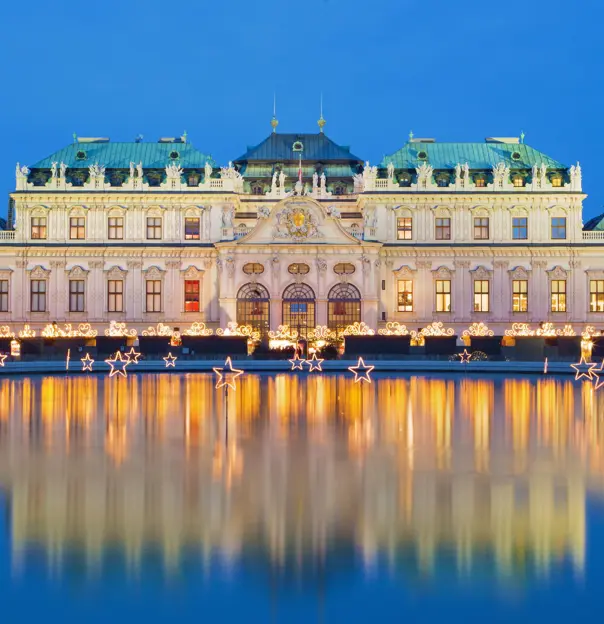 Shot of Schoenbrunn Palace, a wide, beige coloured building with turquoise roofs, in front of a bright blue night sky. The palace has gold, swirly light fixtures along the bottom. In front, is a large pond with lit, stand up stars dotted in.