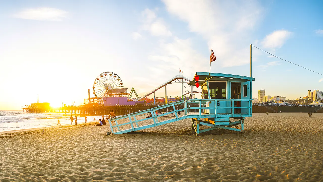 A teal blue lifeguard hut on Santa Monica Beach, with the iconic pier and Ferris wheel in the background as the sun goes down