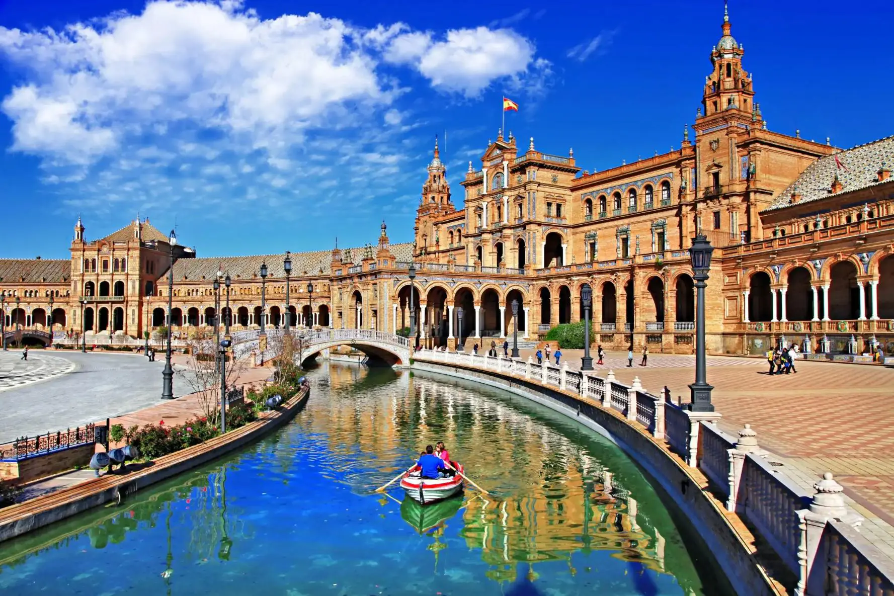 A couple rows a boat along the canal at Plaza de España in Seville, Spain, with the historic buildings and a bright blue sky reflected in the water