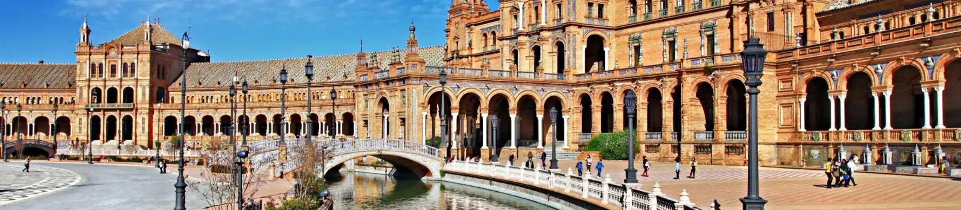 A couple rows a boat along the canal at Plaza de España in Seville, Spain, with the historic buildings and a bright blue sky reflected in the water