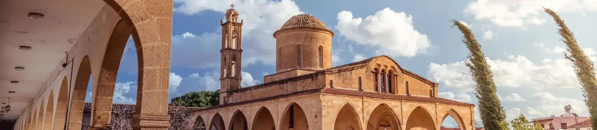 A stone Greek Orthodox monastery with arched walkways, a domed roof, and a bell tower. A well-kept garden with flowers and shrubs lines the foreground under a sunny sky