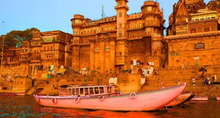 The ghats of Varanasi seen from the River Ganges, with historic sandstone palaces and colourful boats reflecting in the calm evening water