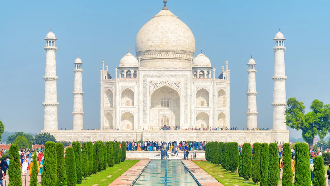 A symmetrical view of the Taj Mahal on a clear day, showing its white marble façade, ornate minarets, and carefully trimmed topiary gardens