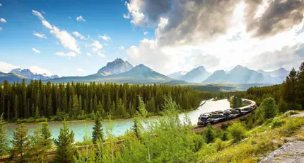 The Rocky Mountaineer train travelling through the Canadian Rockies, with rugged mountains in the background and dense evergreen forests lining the tracks