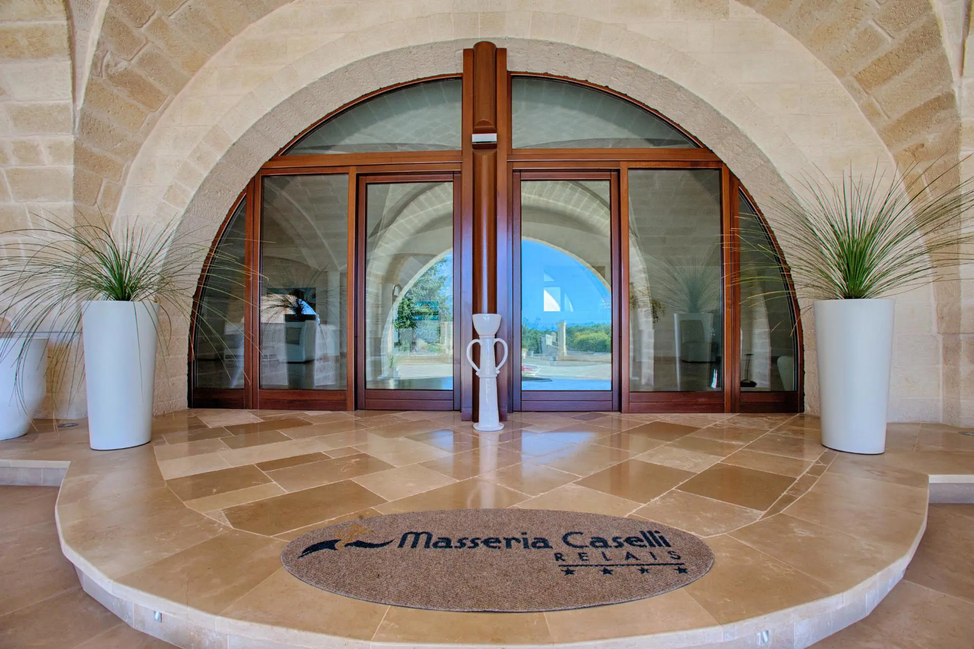 Entrance to Masseria Caselli Hotel, Italy, featuring arched stone architecture, large glass doors, and modern planters