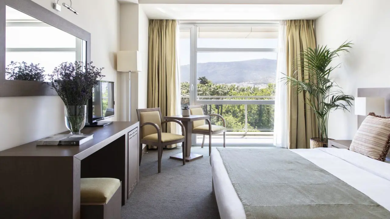 A bright hotel room at Amalia Hotel, Olympia with modern furnishings, a large bed, desk, and balcony view of greenery and hills
